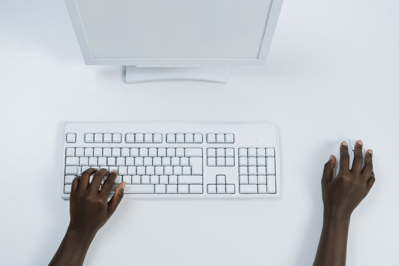 Top view of a minimalist workspace with a computer and keyboard, featuring dark-skinned hands using mouse and keyboard.